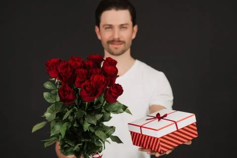 Young man is posing on grey background with gift box and red roses in hands Stock Photos