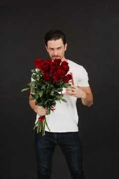 Young man is posing on grey background with gift box and red roses in hands Stock Photos