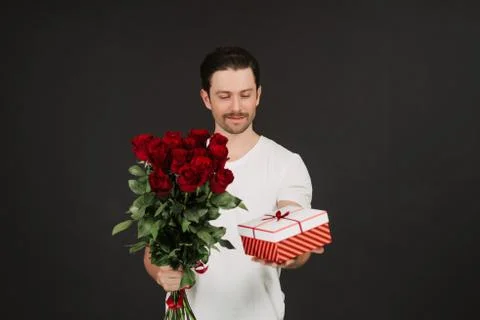 Young man is posing on grey background with gift box and red roses in hands Stock Photos