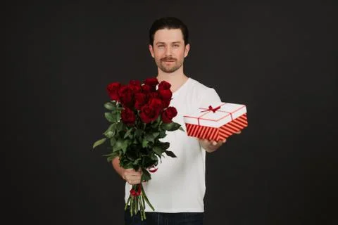 Young man is posing on grey background with gift box and red roses in hands Stock Photos