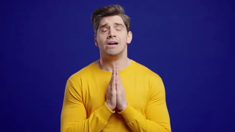 Young Man Praying With Fingers On Blue Background. Guy Begs Satisfy Desires Stock Footage 321096764