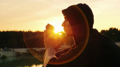 Young man praying at sunset. He sits in profile to the camera hood, clasped his Stock Footage 66113093