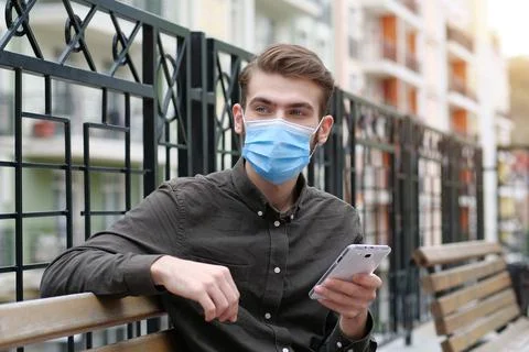 Young man in protective face mask sits on bench and holds in hand smartphone Stock-Fotos