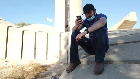 Young man with protective mask checks his mobile in a public park, a sunny day. Stock Footage 135428974