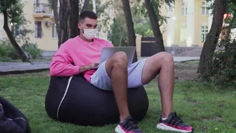 Young man with protective mask sitting in a bag chair in the park and working on Stock Footage 143459567