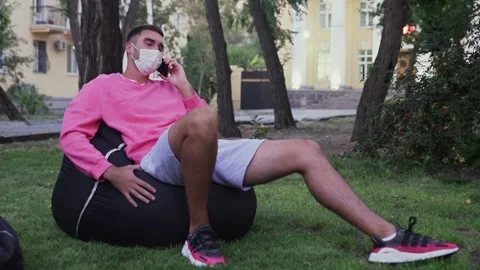 Young man with protective mask sitting in a bag chair in the park and talking on Stock Footage 143459915