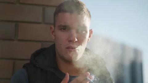 young man puffing cigarette smoke with thoughtful look in urban background