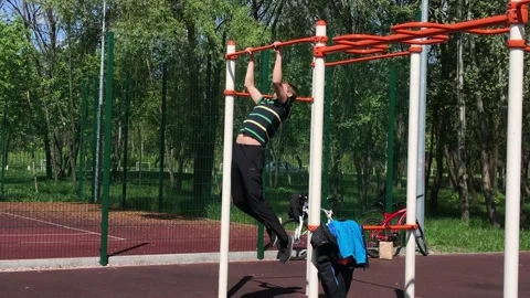 A young man pulls himself up on horizontal bars against a green park Stock Footage 130491551