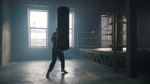 Young man punch the bag hard near boxing ring in industrial interior silhouette Stock Footage 91587619