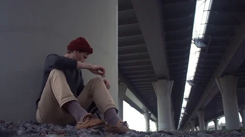 Young man puts on a blue jacket sitting on the ground under road bridge Stock Footage 80285832