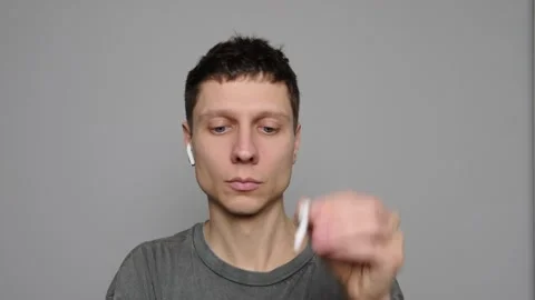 A young man puts wireless Bluetooth earbuds into his ears, gray background. Musi Stock Footage 296884000