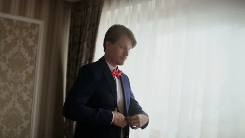 Young man putting on jacket standing by window. Groom is preparing for his Stock Footage 105838685