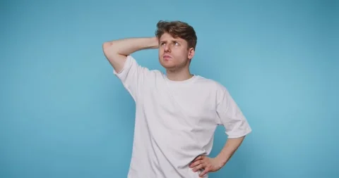 Young man with puzzled expression stands against blue background while thin.. Stock Footage 316180791