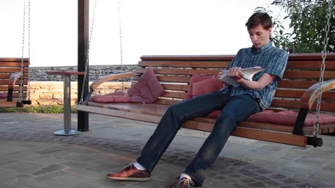 Young man reading and relaxing on a gazebo swing on a summer evening Stock Footage 76839363