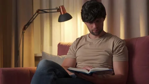 Young man reading and studying a book while sitting on the couch at home in the Stock Footage 211638113