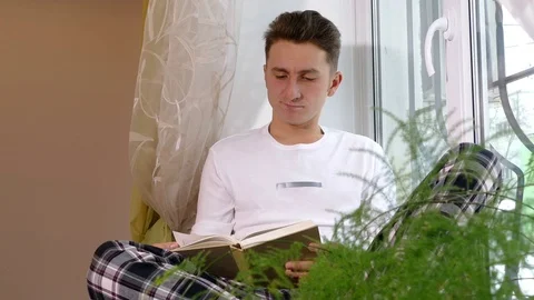 Young man reading book and sitting on the windowsill Stock Footage 71779241