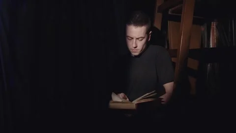 Young man is reading a book in a dimly lit room. The light from the lamp cast Stock Footage 344914074