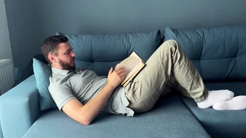 A young man is reading a book lying on the couch. The concept of home recreation Stock Footage 299730854