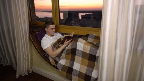 A young man is reading a book. A man lies on a window sill by the window with a Stock Footage 126388723