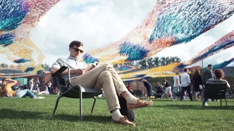 Young man reading a book sitting on chair at city park. Summer day Stock Footage 79356570