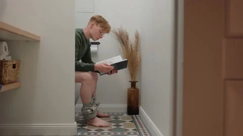 Young man reading a book sitting in white modern toilet 스톡 동영상 164381326