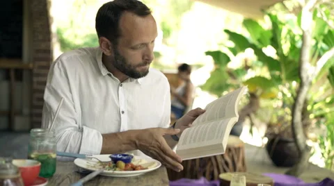 Young man reading book while eating lunch in cafe in the garden, 4K Stock Footage 68925447