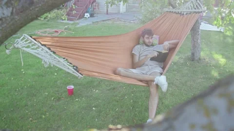 A young man reading book while lying in a hammock in the park Stock Footage 143484764