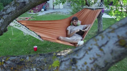 A young man reading book while lying in a hammock in the back yard Stock Footage 143485273
