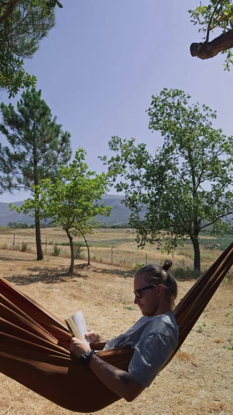 Young man reading a book while relaxing in a hammock Видео 343658980