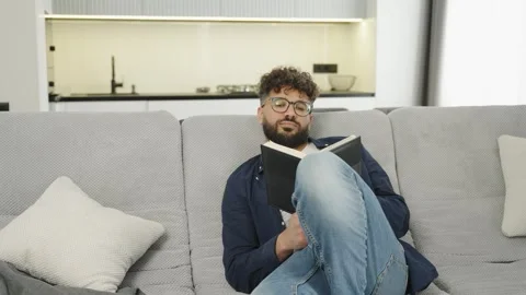 Young man reading book while relaxing on living room couch Stock Footage 345128399