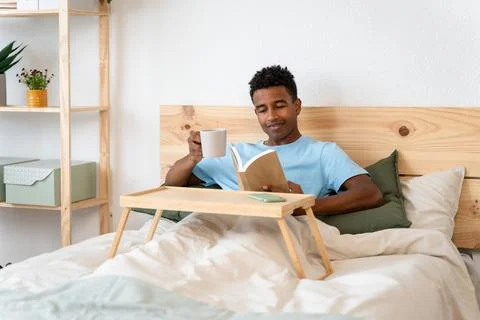 Young man reading a book while enjoying a cup of coffee in bed. Stock Photos