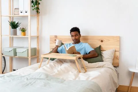 Young man reading a book while enjoying a cup of coffee in bed. Stock Photos