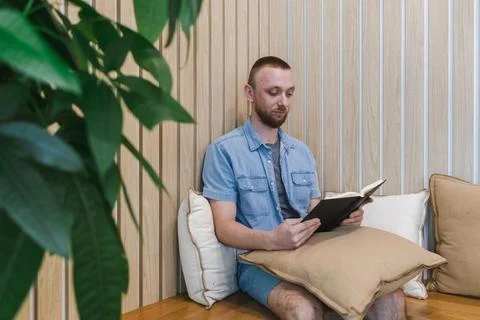 Young man reading a book while sitting on a cushion in a cozy corner during.. 库存照片
