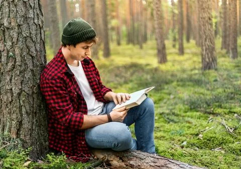 Young man reading a book while relaxing near a tree in a pine forest. Conce.. Stock Photos