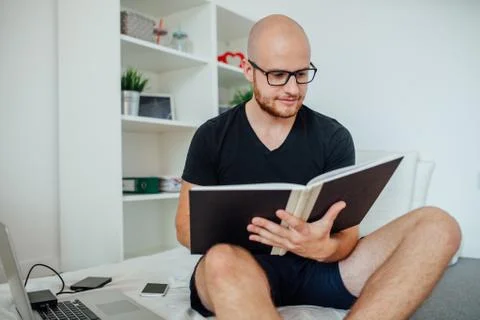 Young man is reading something from notepad and sitting on the bed. Home back Stock Photos