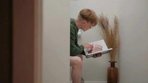 Young man reading a textbook sitting in white modern toilet Video stock 164380439