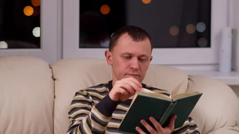 A young man reads a book while sitting on a sofa in the evening Stock Footage 132355639