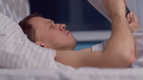 Young man reads a book while lying on his back, at night in bed, camera movement Stock Footage 132639850
