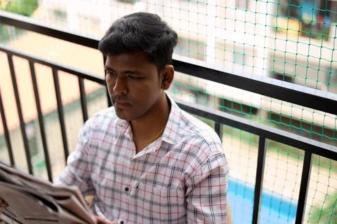 A young man reads the newspaper while seated on a balcony in the morning su.. Stock Photos