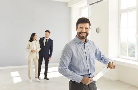 Young man realtor standing in empty new apartment holding contract with a couple Stock Photos