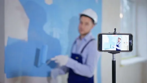 Young man records on smartphone camera how to do finishing work and paints walls 스톡 동영상 146988147