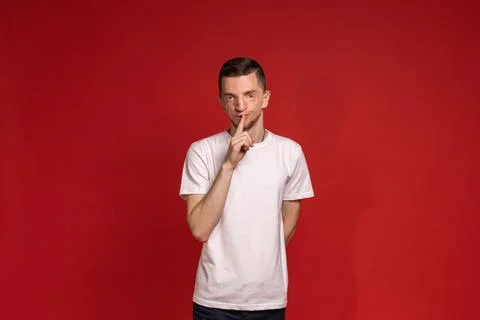 The young man in the red background in the white T-shirt puts his finger to.. Stock Photos