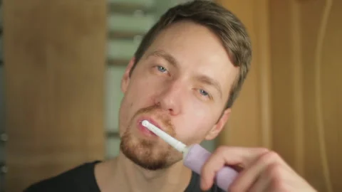 A young man with red beard is brushing his teeth with a white electric brush Stock Footage 265469597