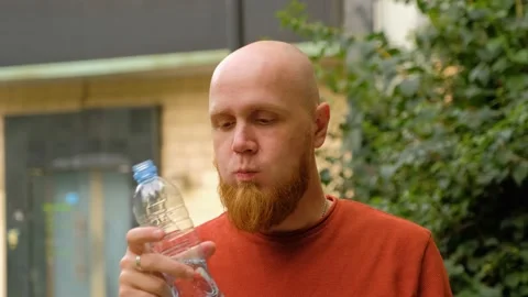 A young man with a red beard drinks water from a plastic bottle on the street. Stock Footage 139111159