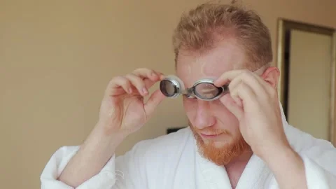 Young man with a red beard in a white robe puts on swimming goggles and leaves Stock Footage 147023279