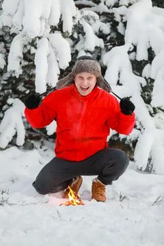 Young man in a red jacket and hat earflaps warms his hands by the campfire in Stock Photos