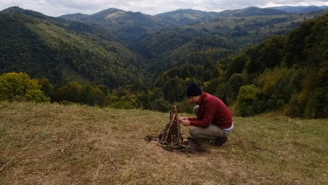 A young man in red jacket kindles a bonfire in mountains. Stock Footage 117051801