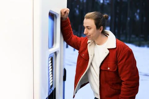 Young man in red jacket leaning on a vending machine in a snowy winter park.. Stock Photos