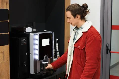 Young man in a red jacket is using an automatic coffee machine in an office.. Stock Photos