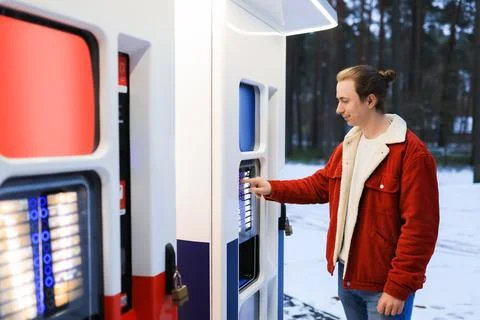 Young man in red jacket using outdoor vending machine in snowy winter park. Stock Photos